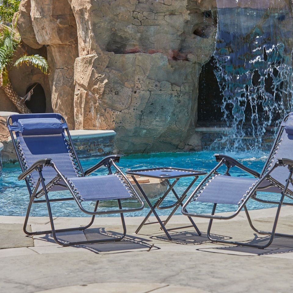 Two navy zero-gravity lounge chairs and a side table beside a pool with a rock waterfall